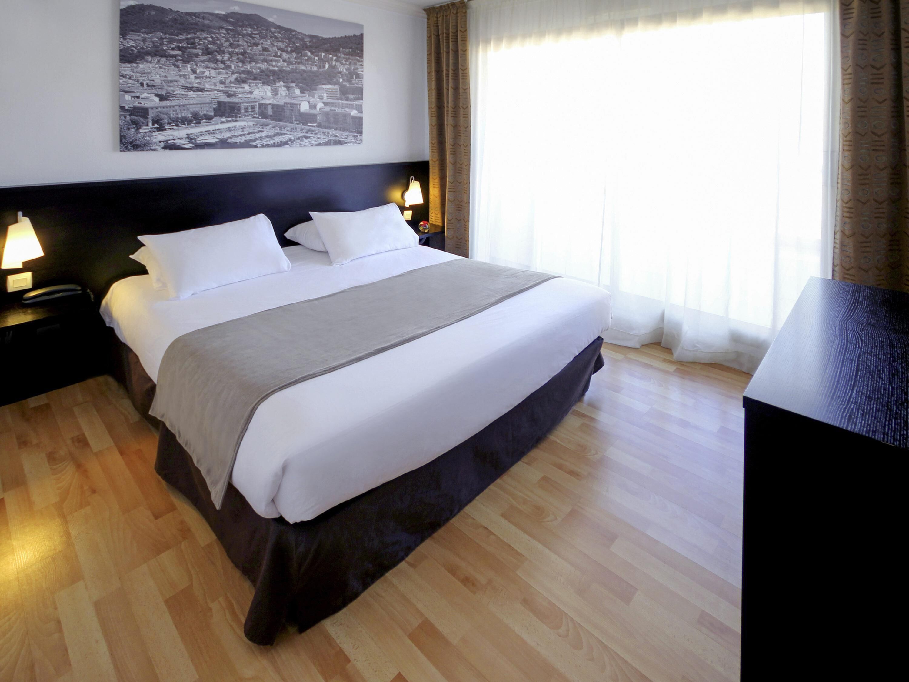 A hotel room with a large bed, a nightstand with a lamp, a wall-mounted picture, and a window with curtains.