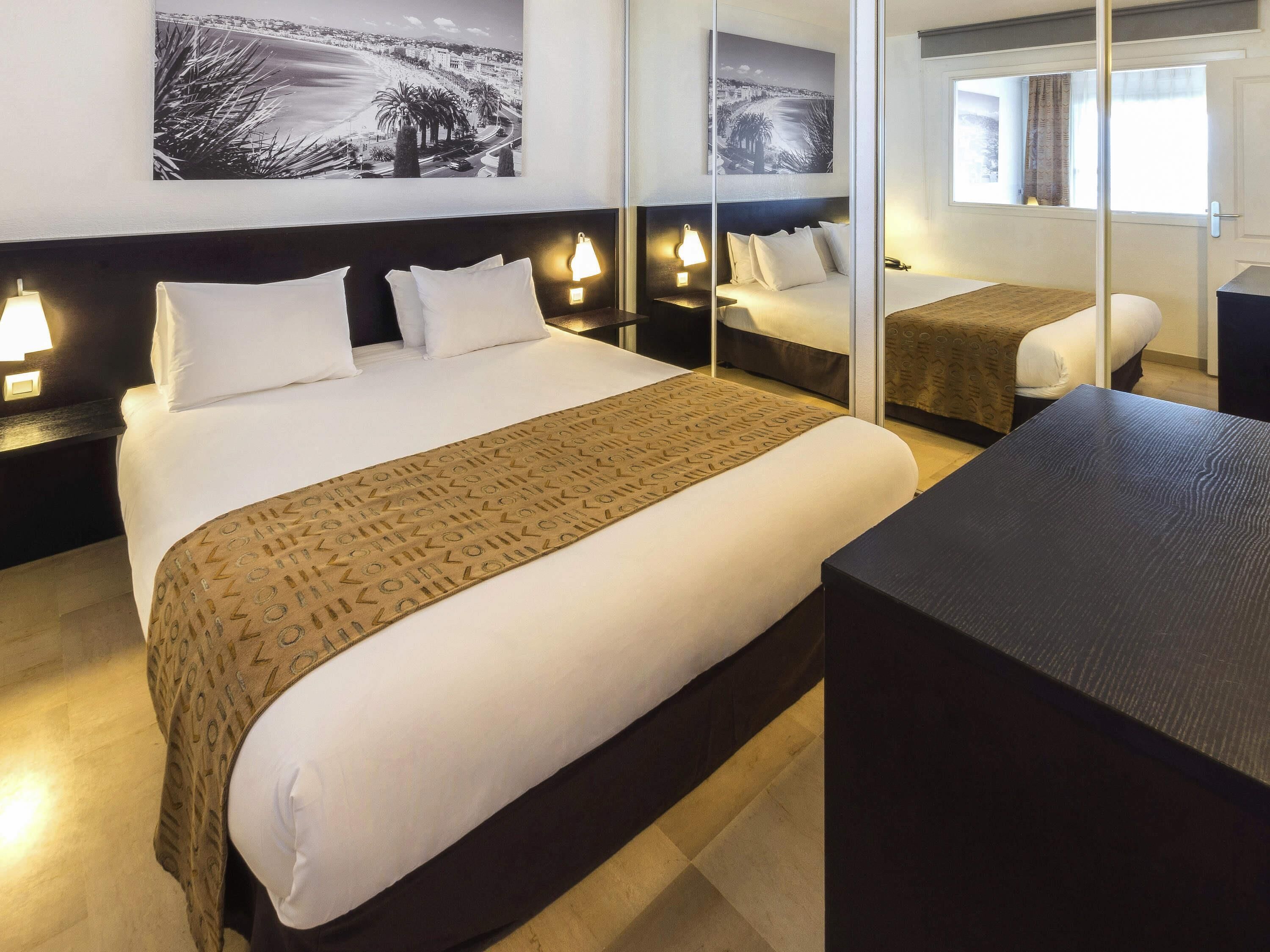 Une chambre d’hôtel avec un grand lit, des tables de chevet avec des lampes, un miroir sur la table de chevet, un petit bureau et une fenêtre avec des rideaux.