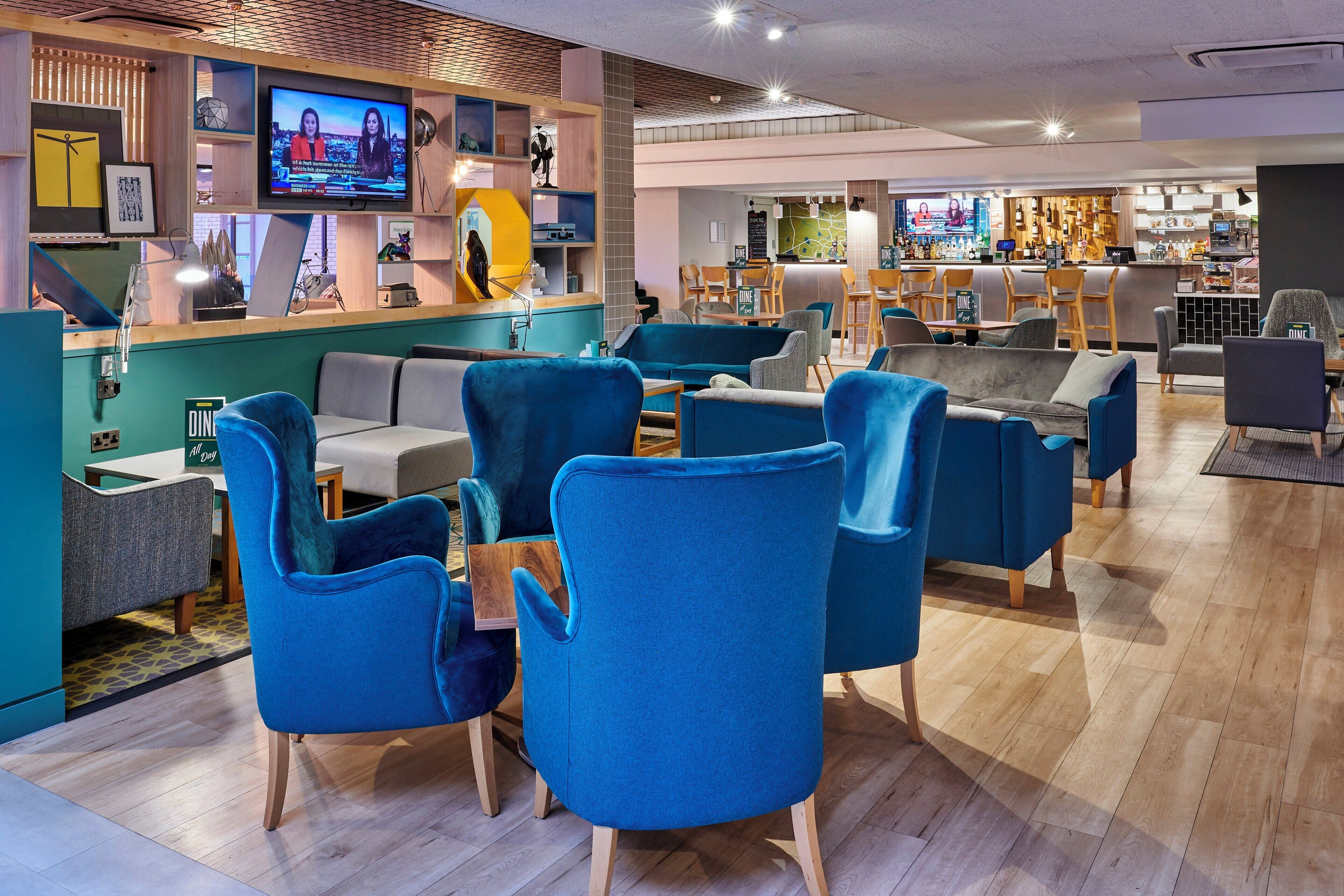 Un área de descanso moderna con asientos tapizados en azul, una zona de bar con televisor y suelo de madera.
