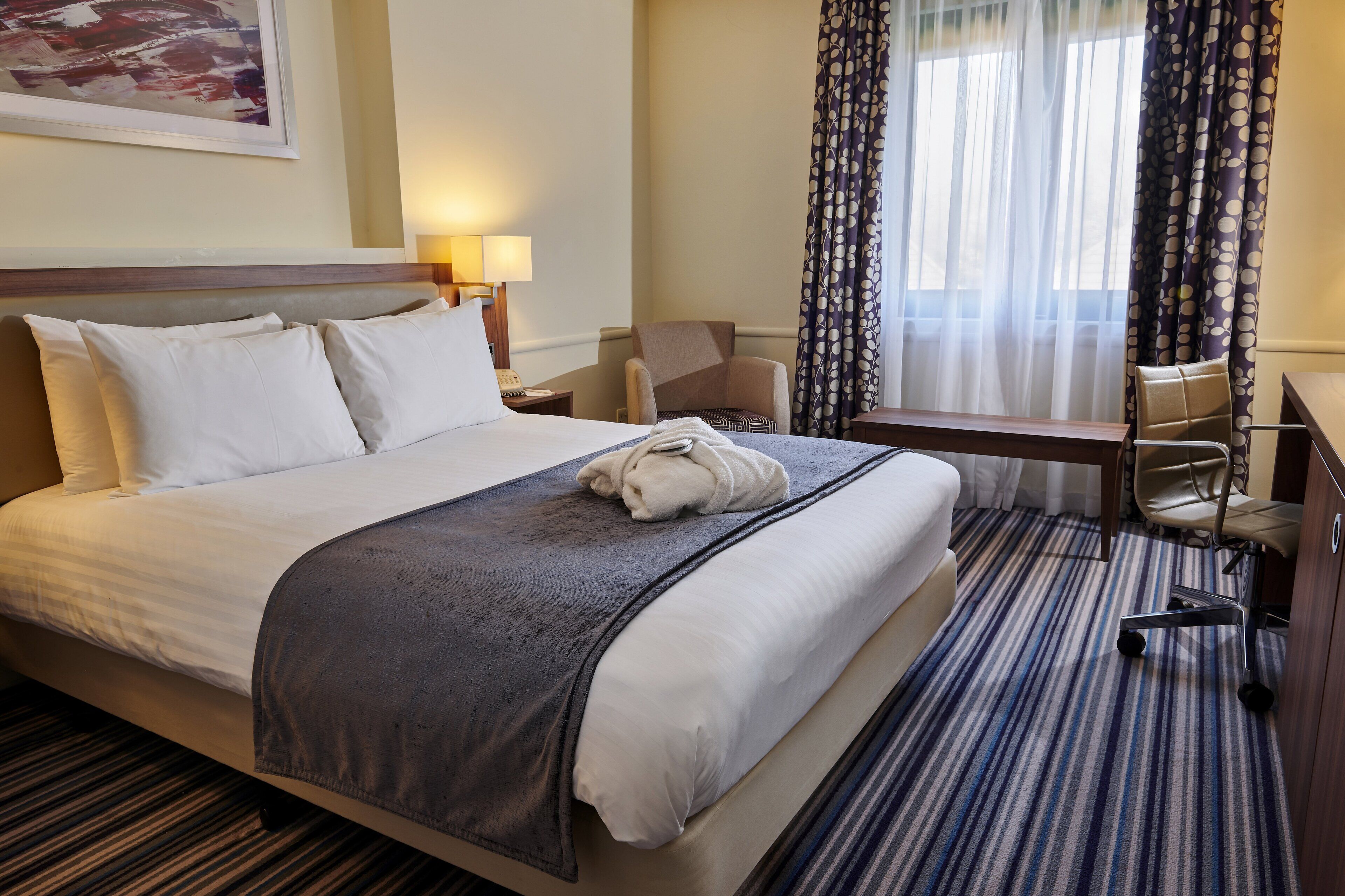 A hotel room with a large bed, a desk, a chair, and a window with curtains.
