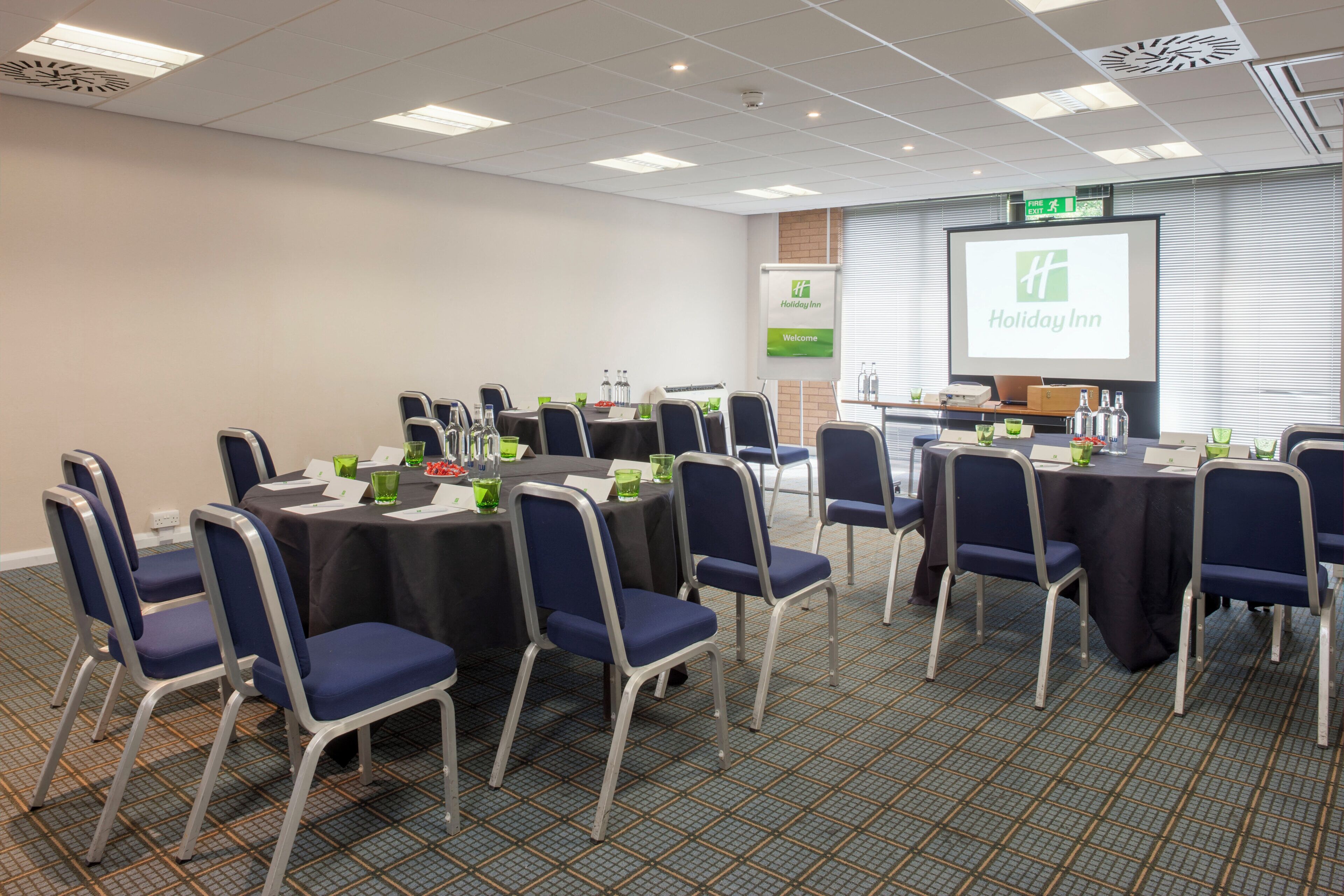A conference room with tables, chairs, a projector screen displaying a Holiday Inn logo, and a welcome sign.