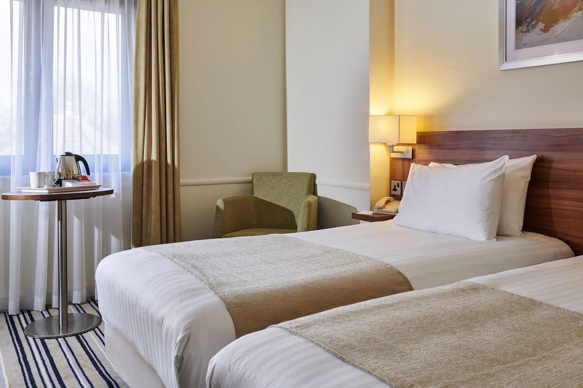 Ein Hotelzimmer mit zwei Betten, einem kleinen Tisch mit Wasserkocher und Tassen, einem Sessel und einer Lampe.