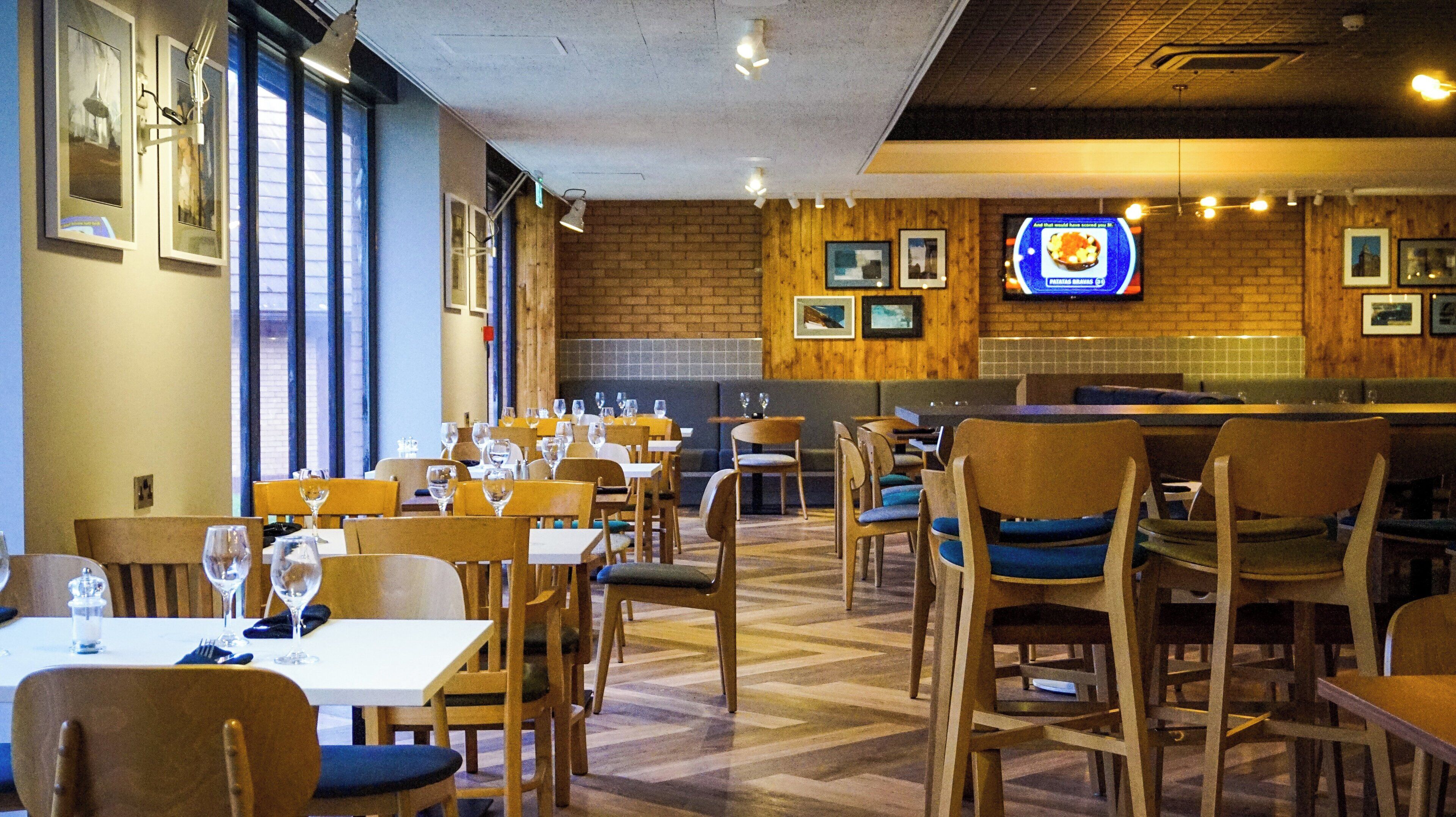 Een restaurantinterieur met houten meubels, tafels gedekt met wijnglazen en een bakstenen muur met ingelijste foto's.