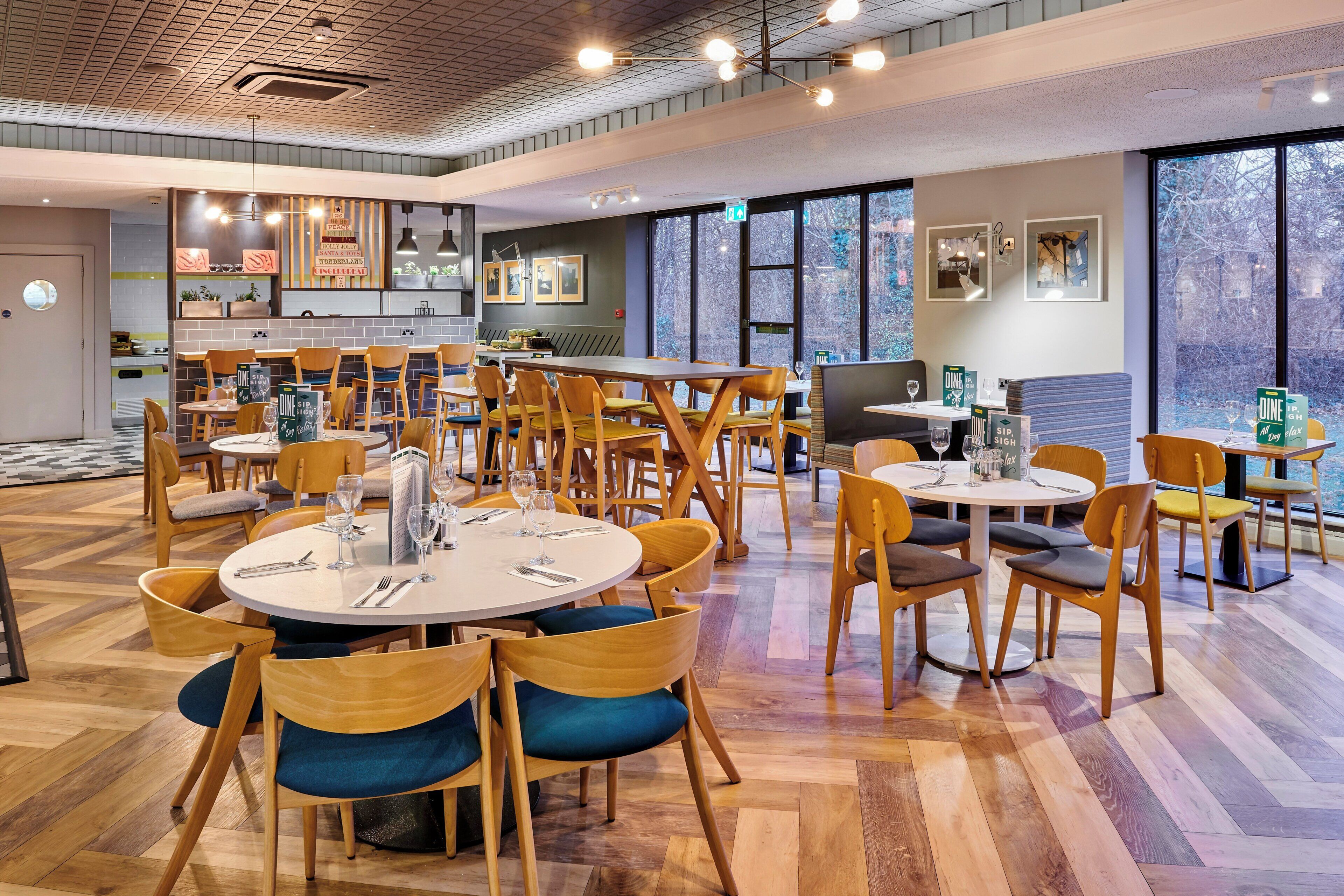 A modern dining area with round tables and chairs, a kitchen in the background, and large windows offering a view of the outdoors.