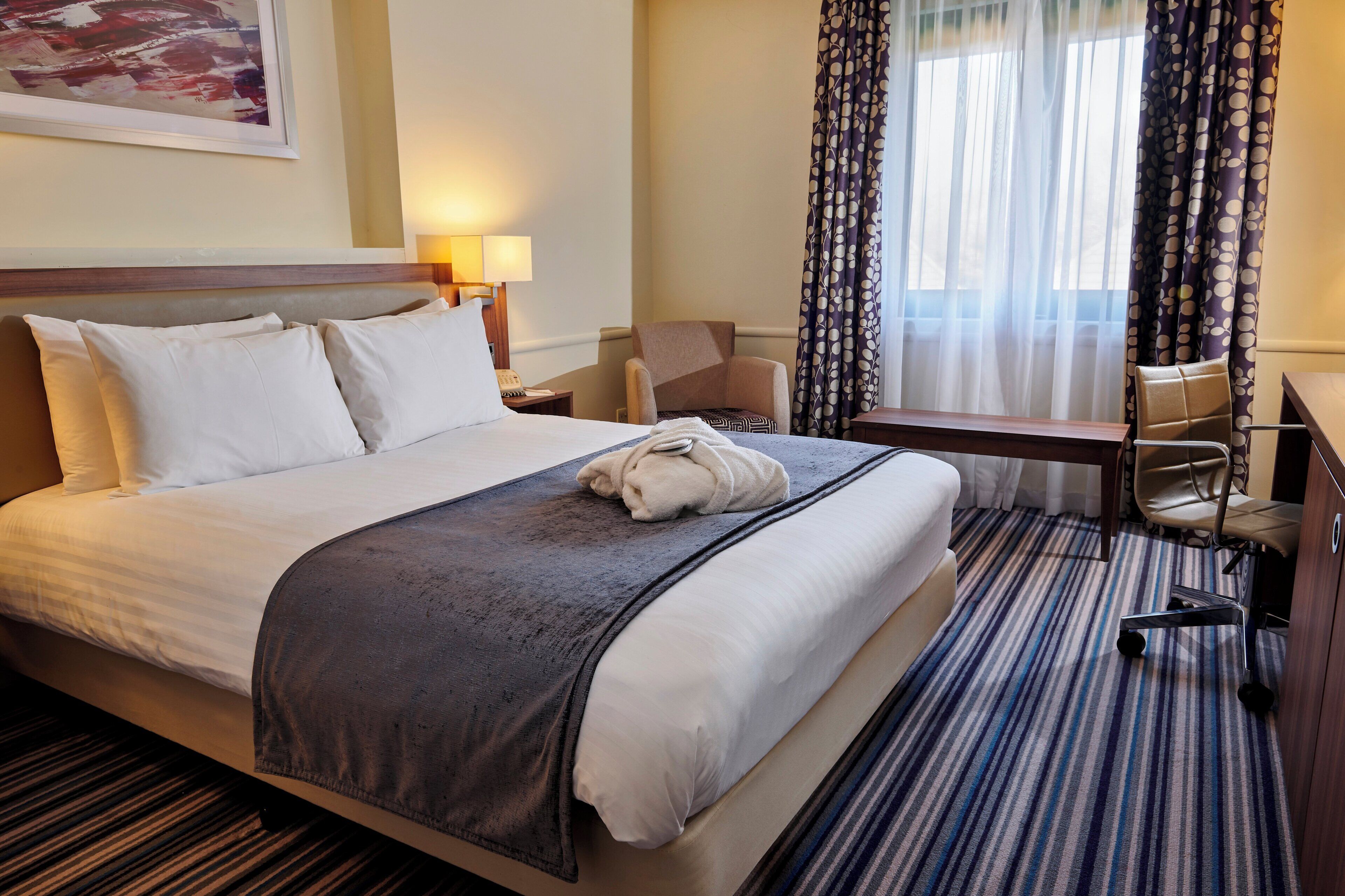 Une chambre d’hôtel avec un lit, un bureau, une chaise, une lampe et une fenêtre avec des rideaux.