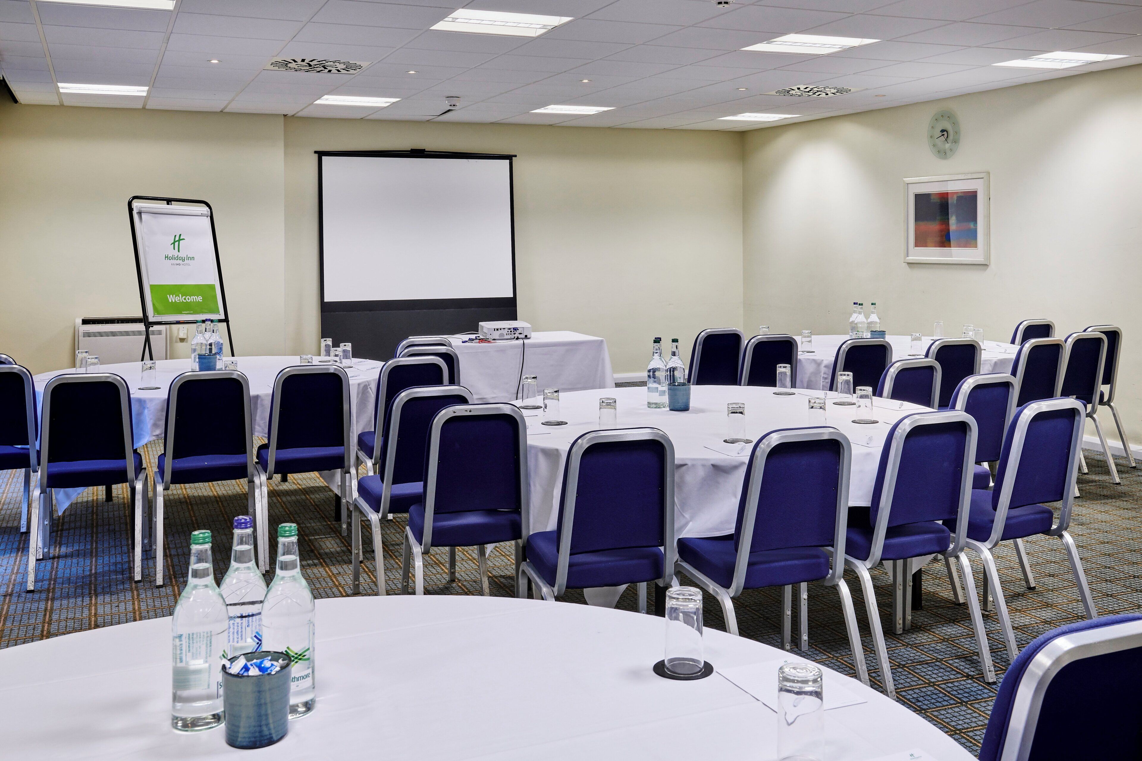 A conference room with round tables, blue chairs, and a projector screen.