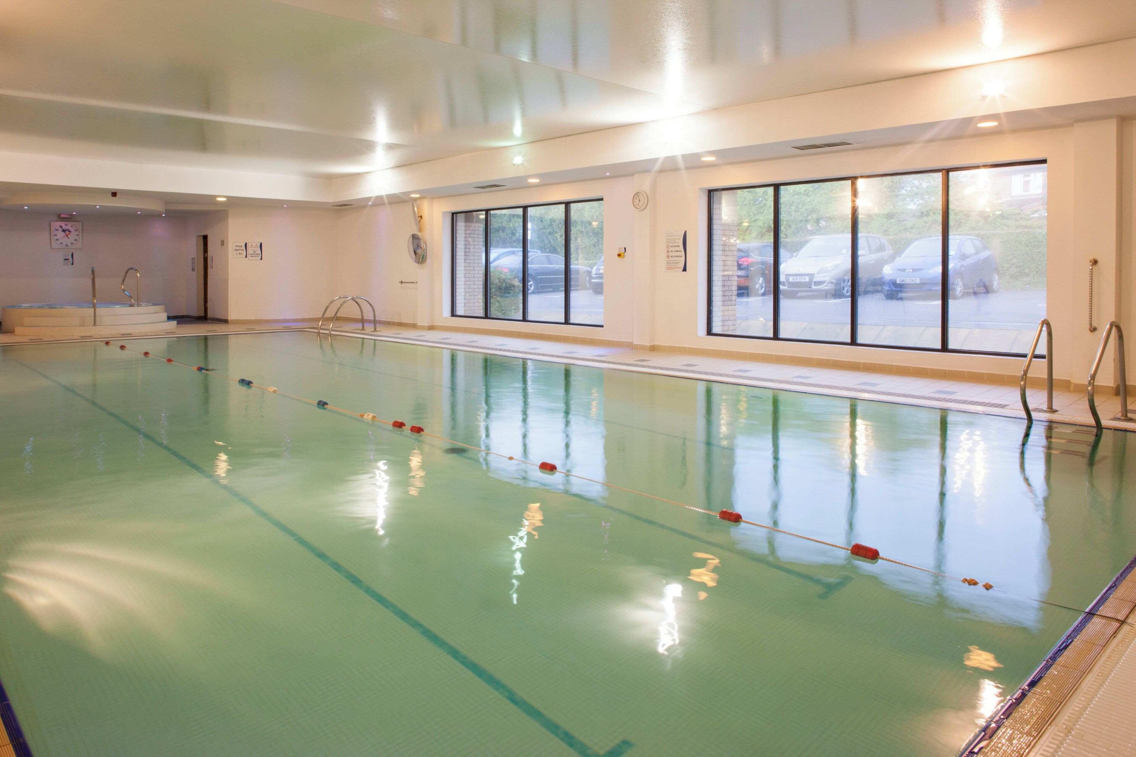An indoor swimming pool with clear lanes, a clock on the wall, and large windows overlooking a parking area.