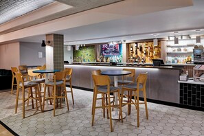 Zona de bar con suelo de baldosas hexagonales, sillas de madera, una barra con pantalla digital y una variedad de botellas y vasos.
