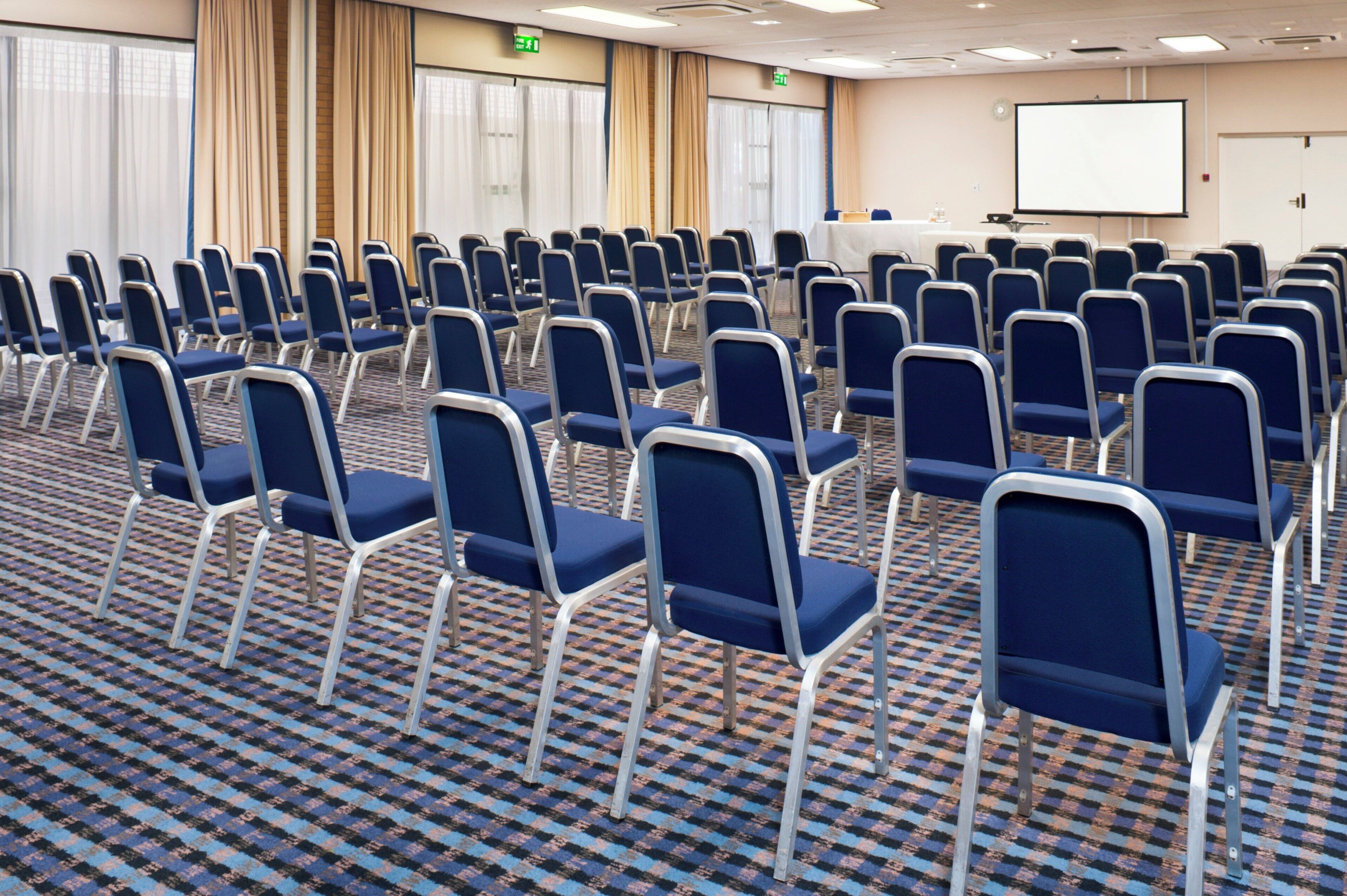 A conference room with rows of blue chairs, a projector screen, and an exit sign.