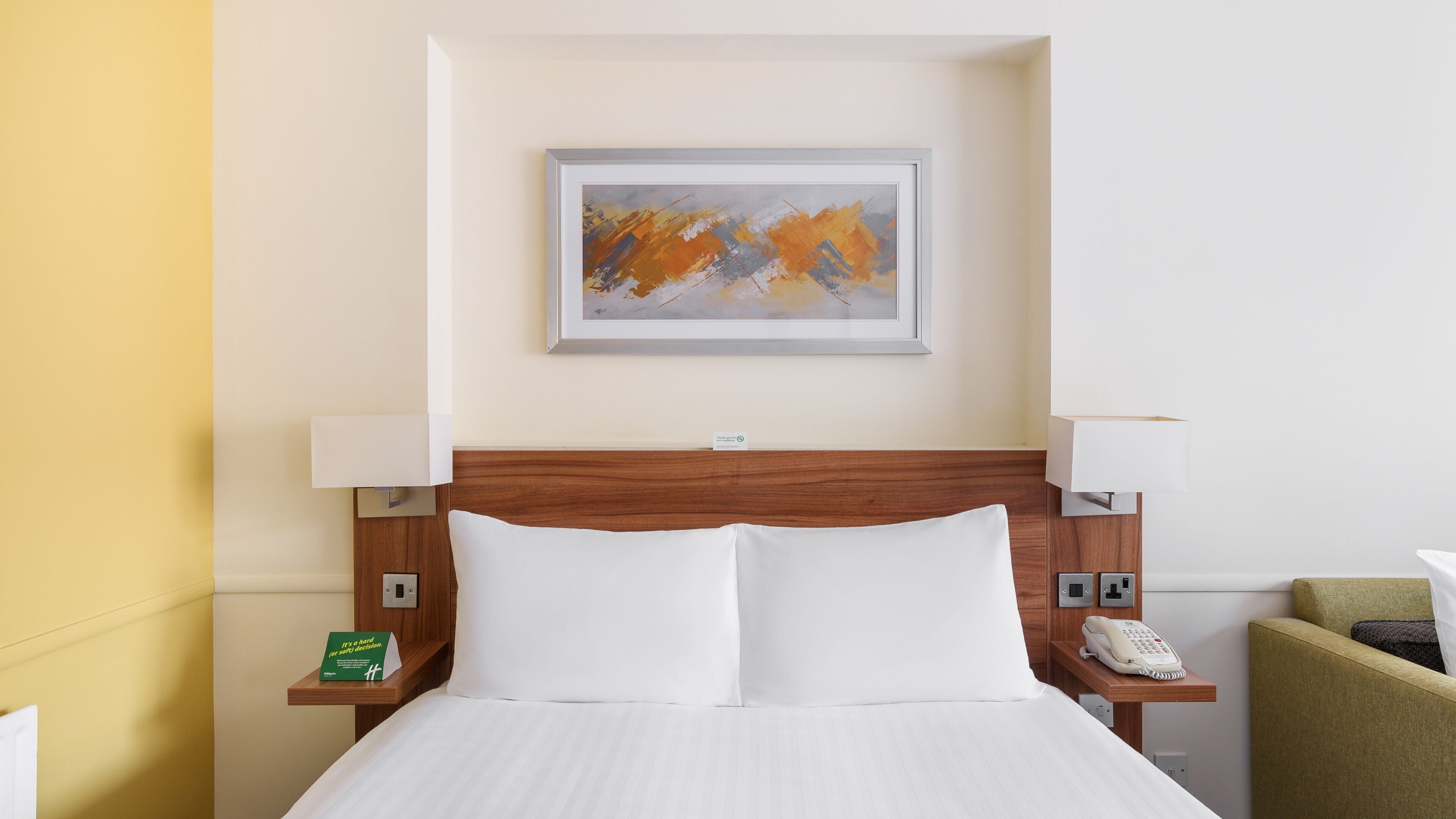 A hotel room with a bed, two bedside tables, a nightstand, a telephone, and a painting on the wall.