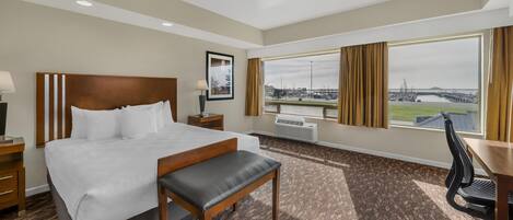 A hotel room with a large bed, a desk, a chair, and a view of a cityscape through the window.