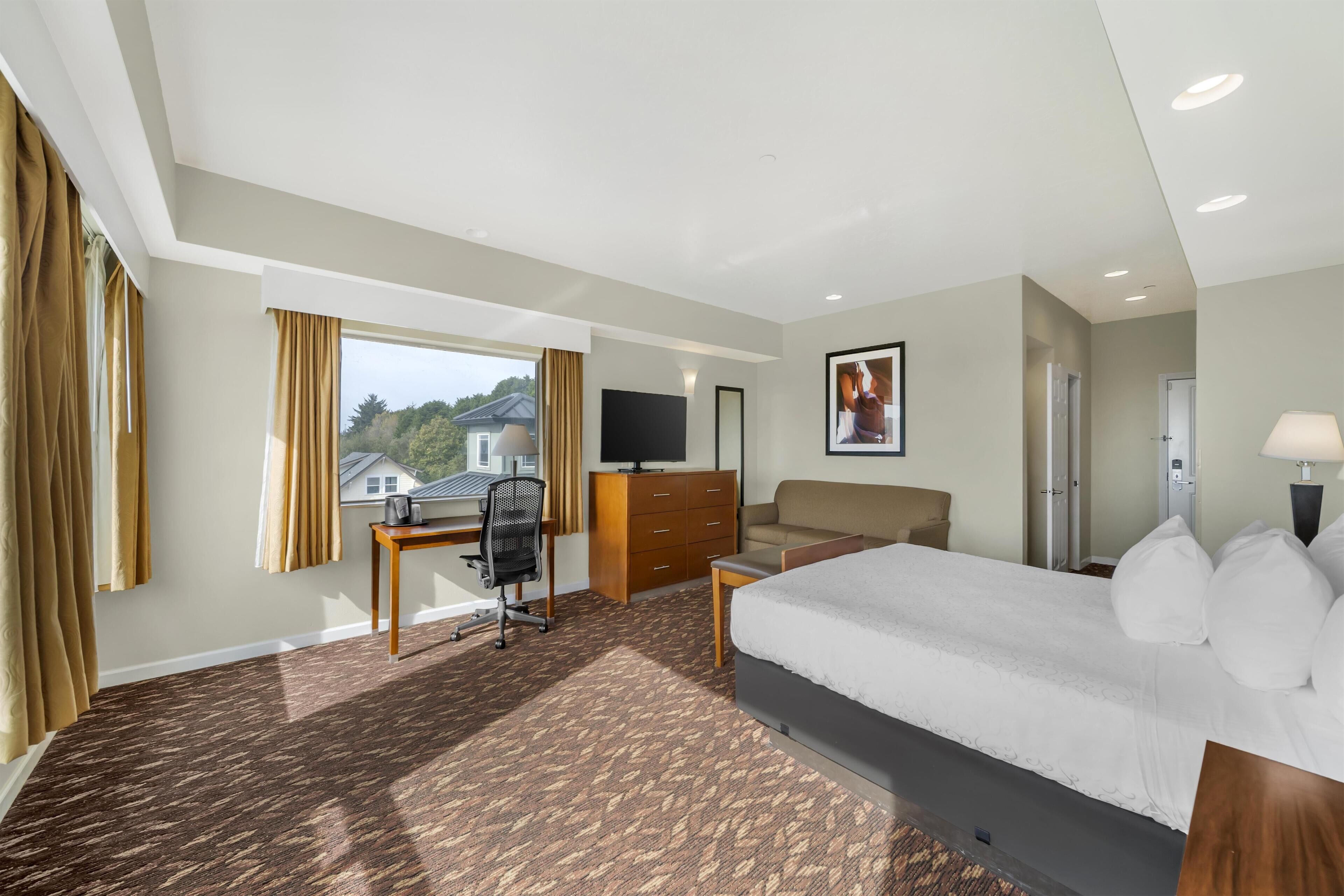 A hotel room with a large bed, a desk with a chair, a dresser, a TV, and a window with curtains.