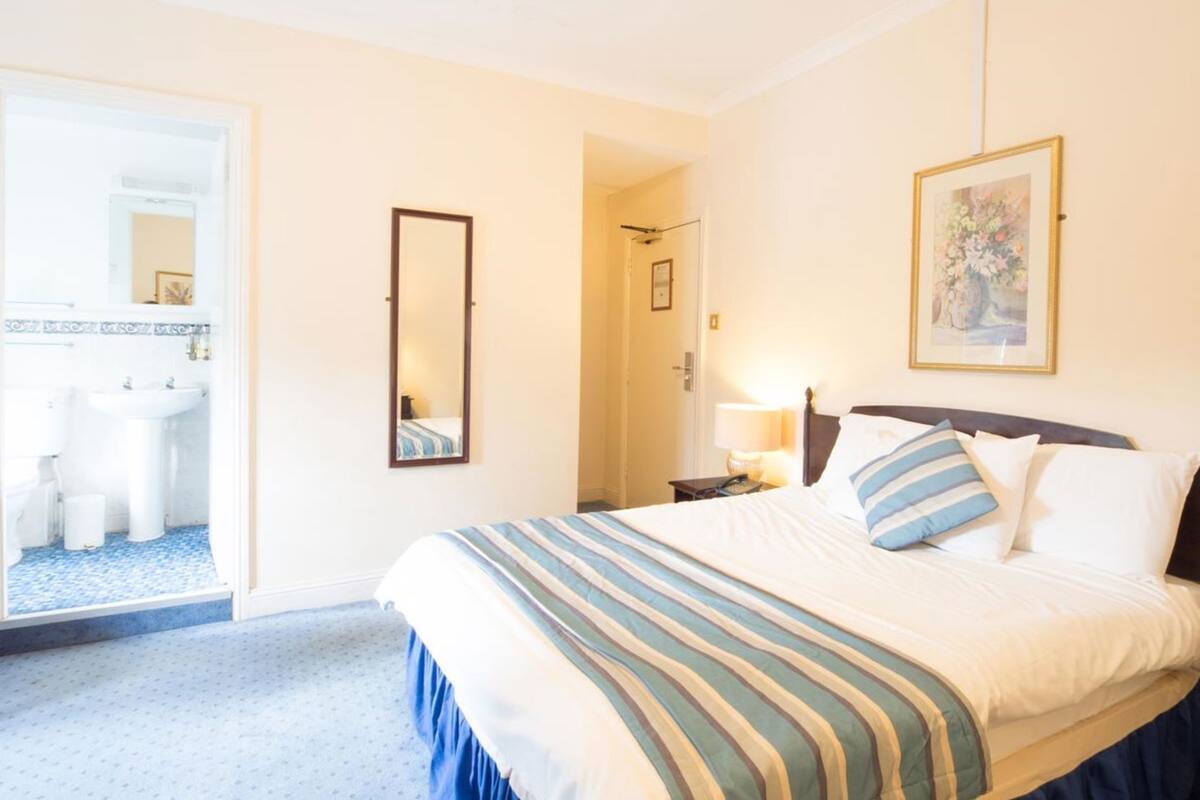 A hotel room with a bed, a bedside table with a lamp, a mirror, a bathroom with a sink and toilet, and a door leading to another room.