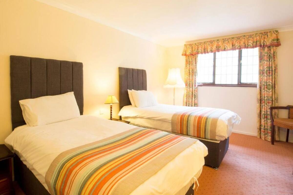 A hotel room with two beds, a window with curtains, a lamp, and a chair.