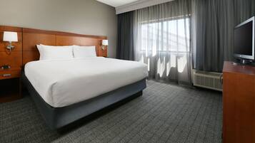 A hotel room with a large bed, two bedside tables with lamps, a flat-screen TV, and a large window with curtains.