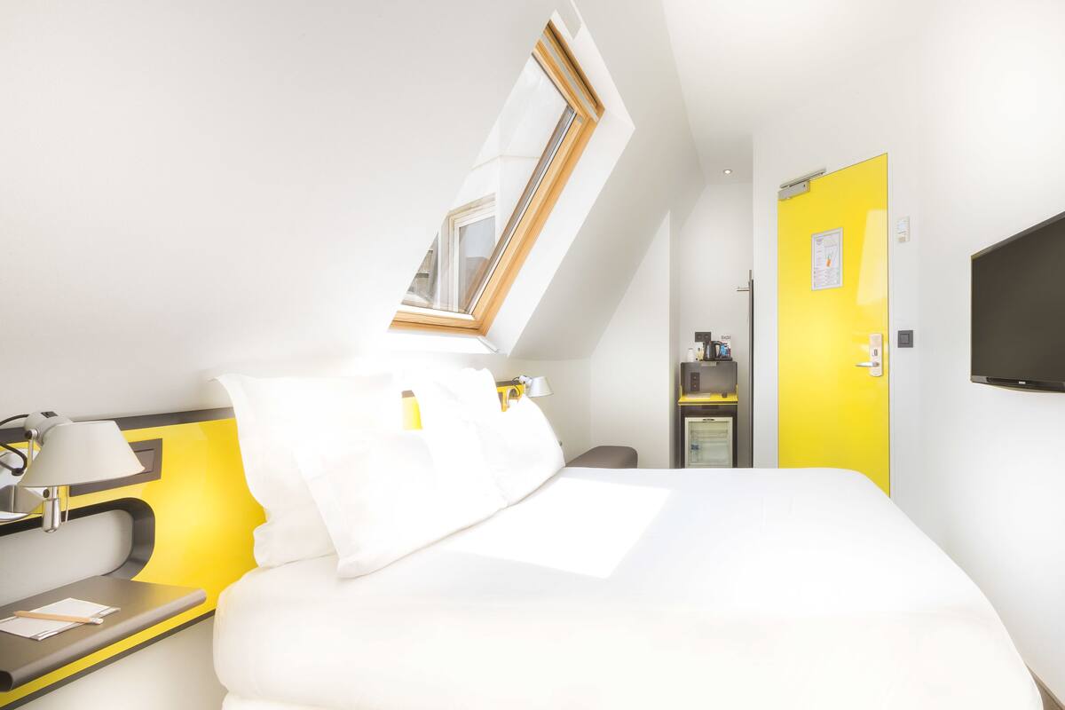 A hotel room with a bed, a desk with a lamp, a television, a yellow door, and a window with a wooden frame.
