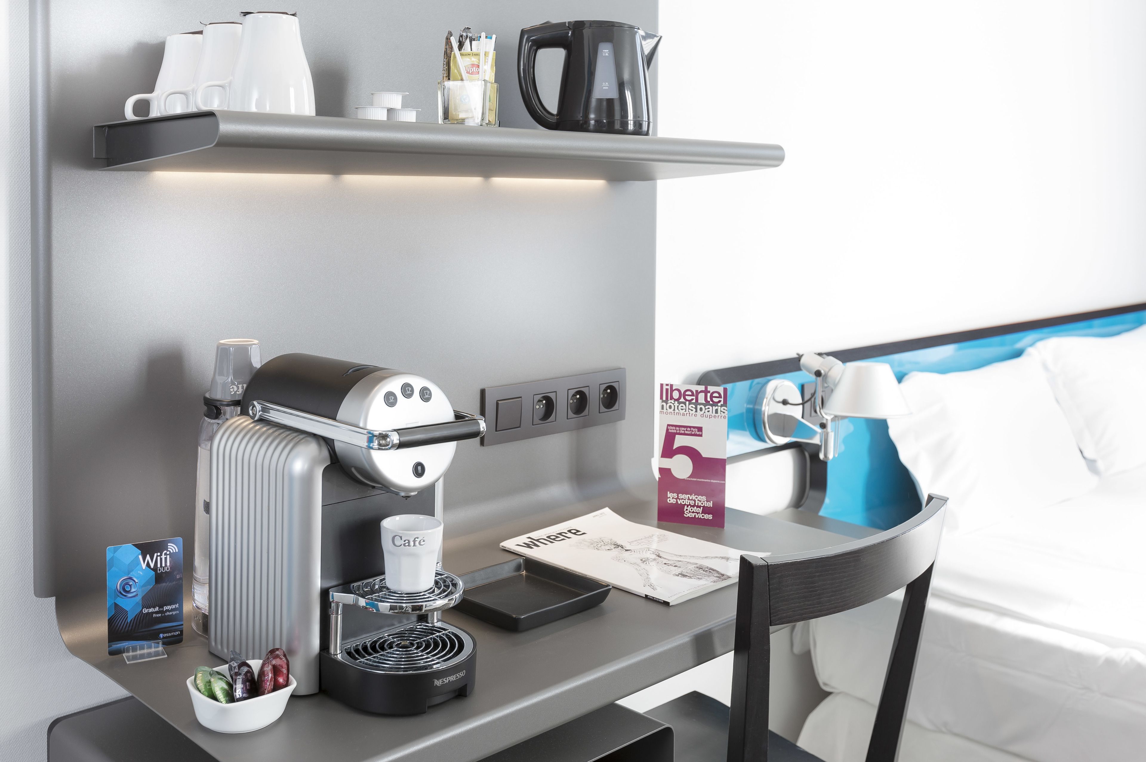A modern hotel room with a built-in coffee machine, a small bowl of fruit, a Wi-Fi sign, and a bed with white bedding.