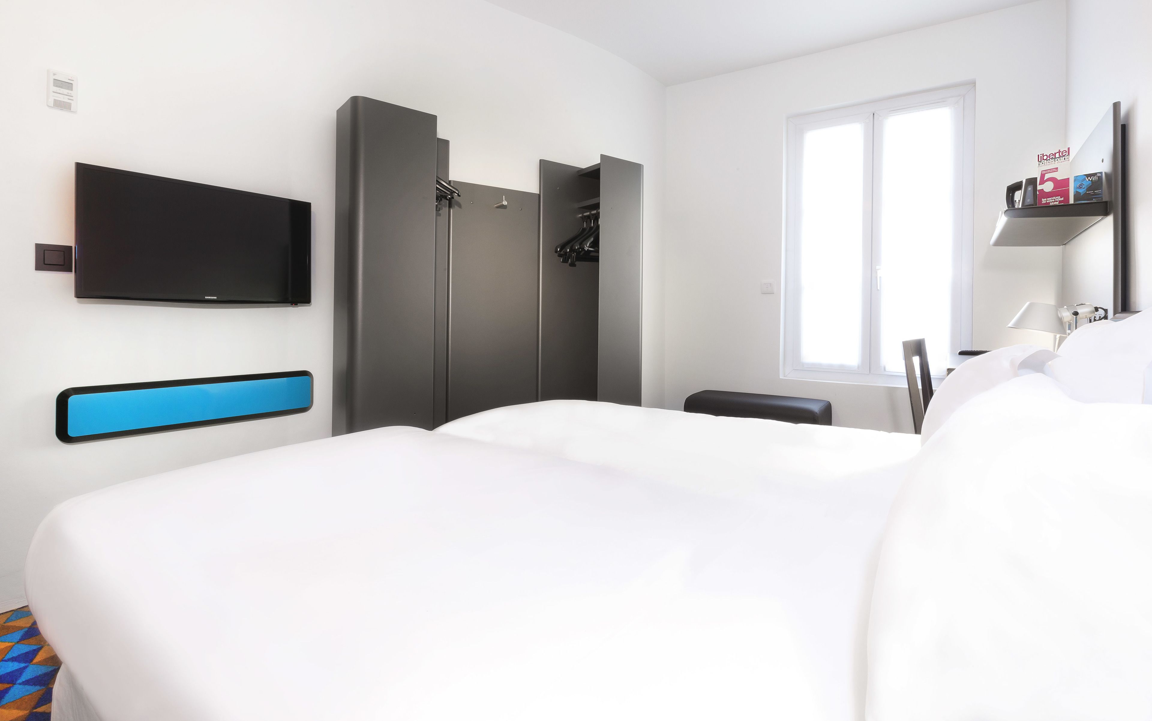 Ein modernes Hotelzimmer mit einem großen Bett, einem wandmontierten Flachbildfernseher, einem Kleiderschrank mit Spiegel und einem Fenster, das natürliches Licht hereinlässt.