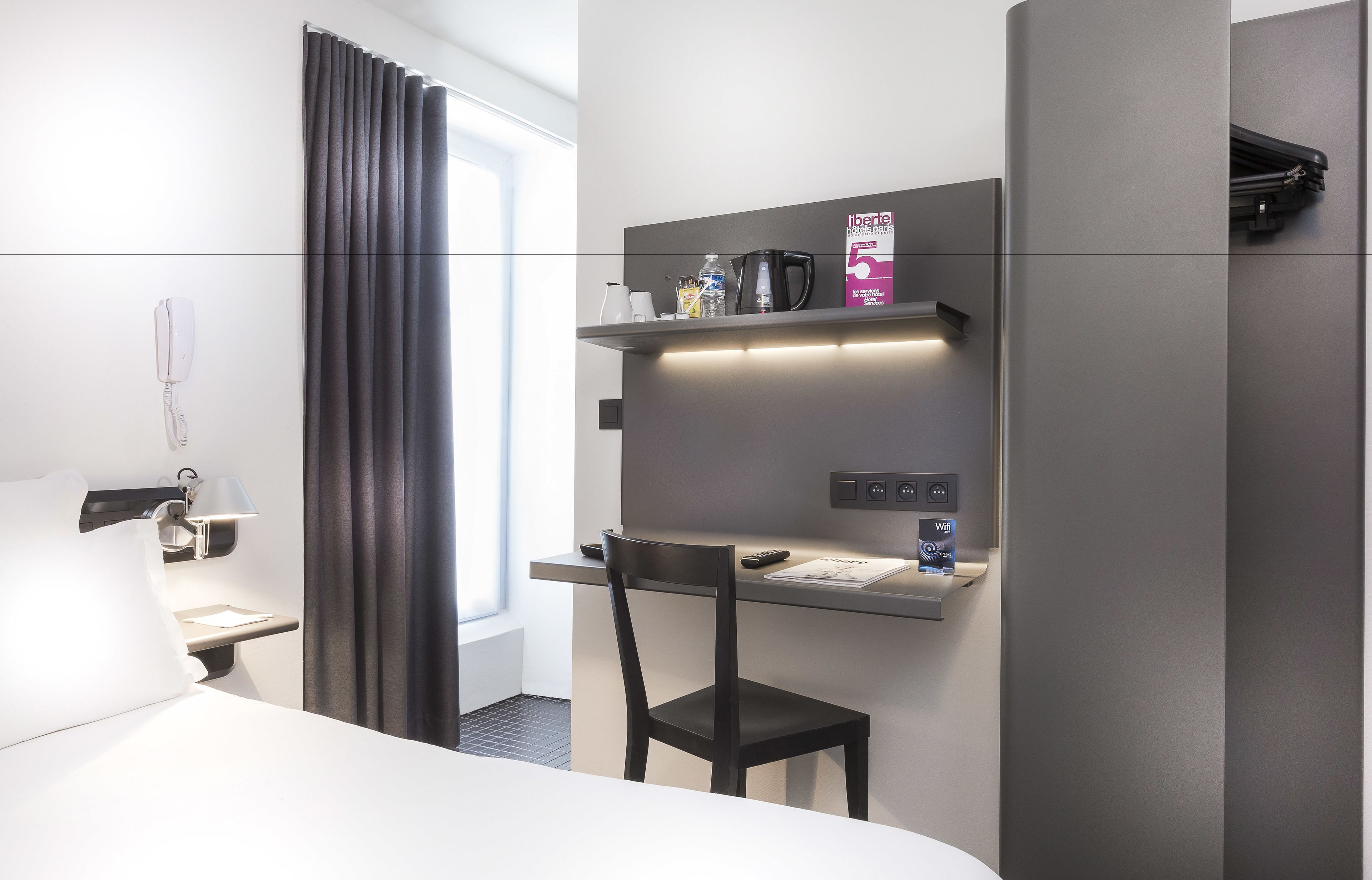 Et moderne hotelværelse med seng, skrivebord med stol, telefon, kaffemaskine og et hyldesæt med toiletartikler.