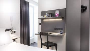 Et moderne hotelværelse med seng, skrivebord med stol, telefon, kaffemaskine og et hyldesæt med toiletartikler.