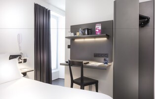 Et moderne hotelværelse med seng, skrivebord med stol, telefon, kaffemaskine og et hyldesæt med toiletartikler.
