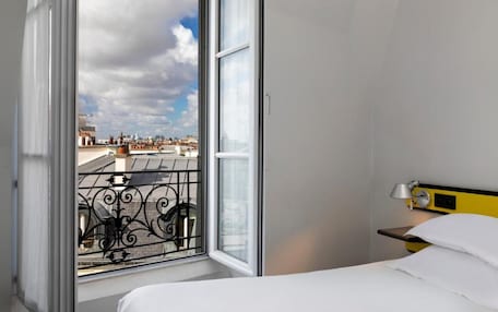 A hotel room with a large window, a bed, a bedside table with a lamp, and a view of the cityscape.