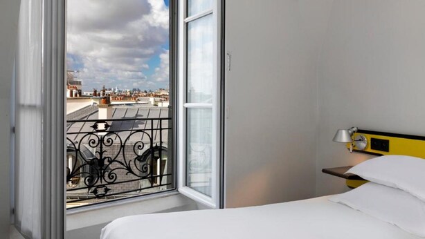 A hotel room with a large window, a bed, a bedside table with a lamp, and a view of the cityscape. - Libertel Montmartre Opéra (Paris)