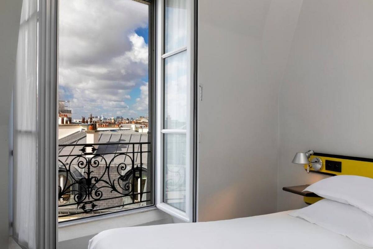 A hotel room with a large window, a bed, a bedside table with a lamp, and a view of the cityscape.