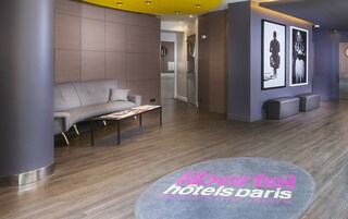 A modern hotel lobby with a grey sofa, a coffee table, and a reception desk. There are framed pictures on the wall and a logo for 'Hotel Paris' on the floor.