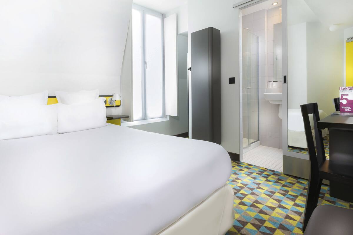 A hotel room with a large bed, a small table with a chair, a bathroom with a glass door, and a patterned floor.
