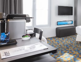 A hotel room with a desk, a chair, a radiator, a TV, a sofa, and a patterned floor.
