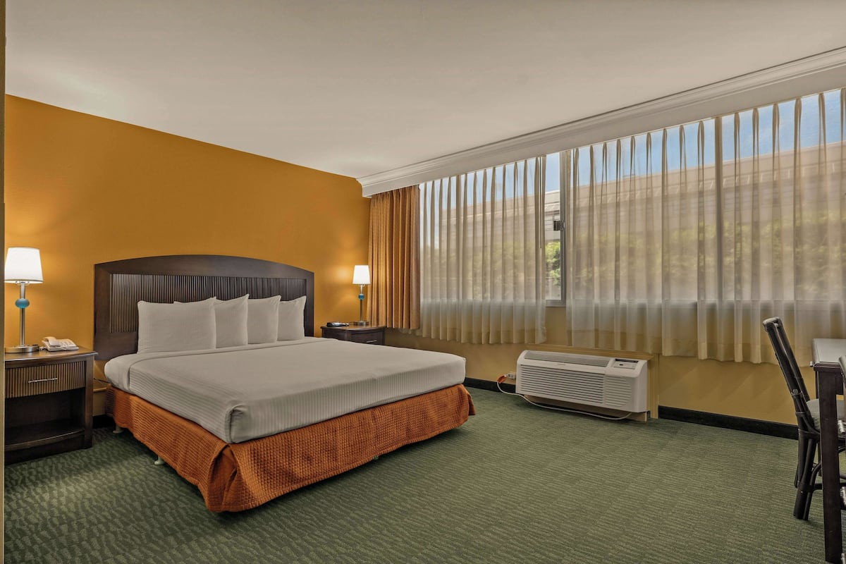 A hotel room with a large bed, two bedside lamps, a desk with a chair, a window with curtains, and an air conditioning unit.