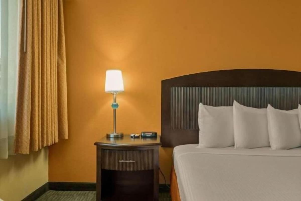 Quarto de hotel com cama, mesa de cabeceira com abajur e janela com cortinas.