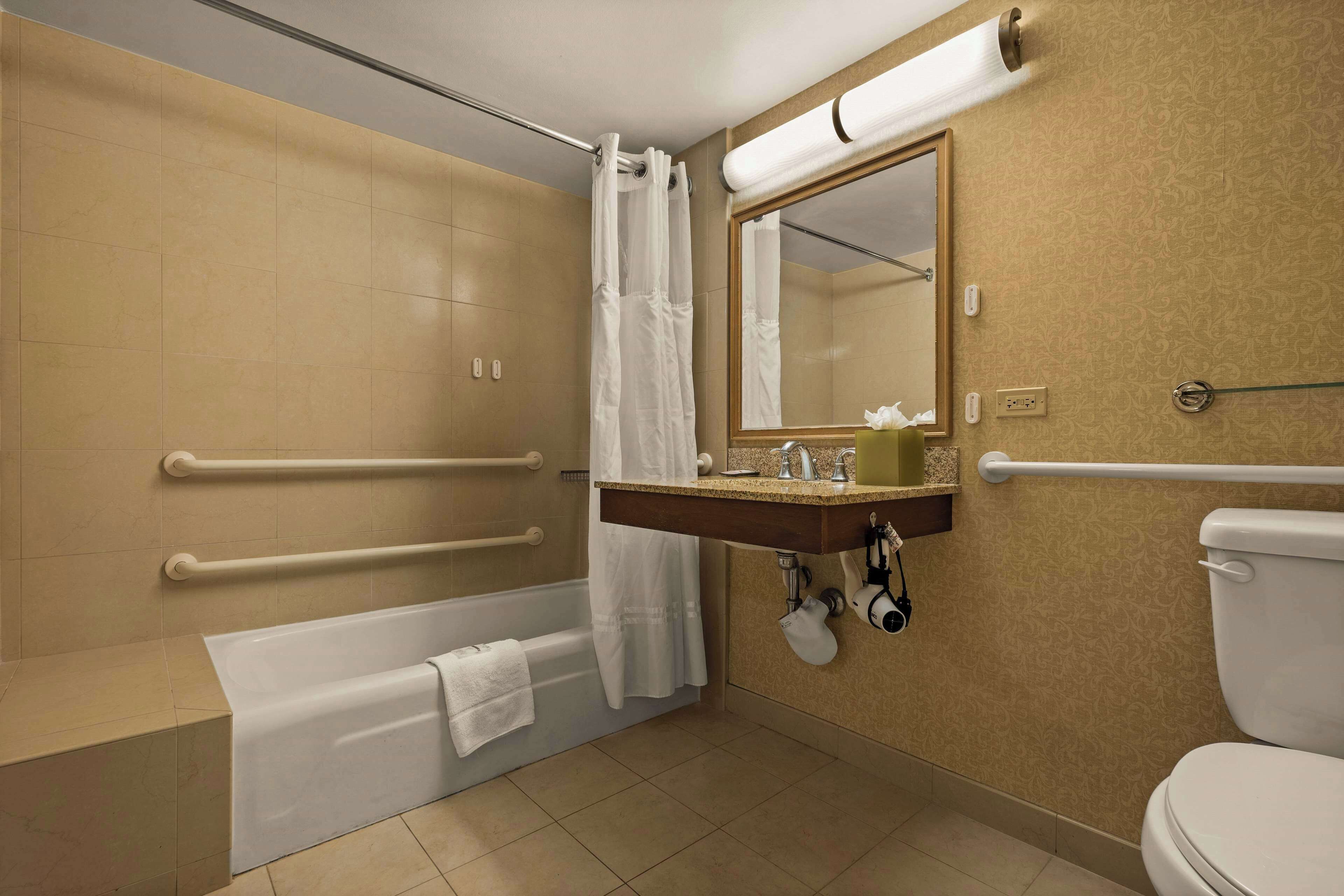 A bathroom with a bathtub, a sink with a mirror, a toilet, and handrails.