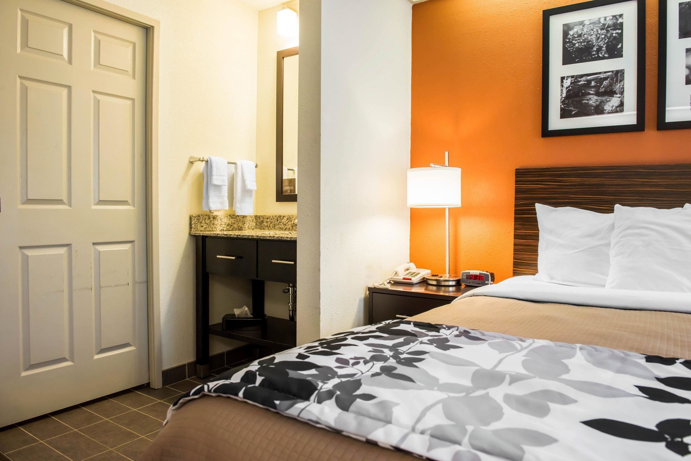A hotel room with a bed, a nightstand with a lamp, a bathroom with a sink and mirror, and framed pictures on the wall.