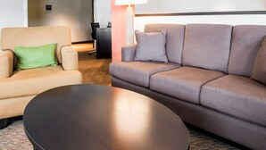 Une chambre d’hôtel avec un fauteuil beige, un canapé gris, une table basse noire et un lampadaire.