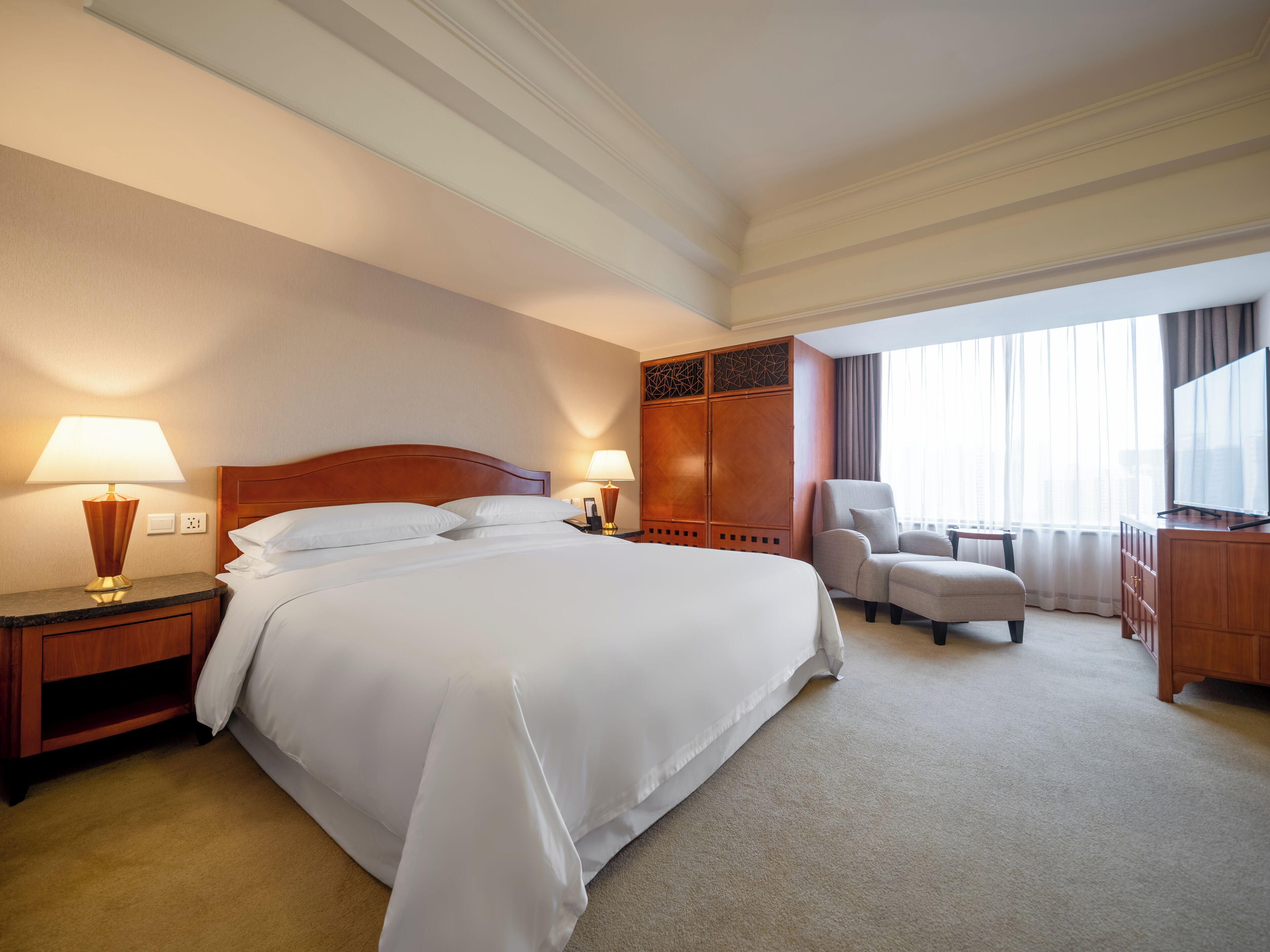 A hotel room with a large bed, two bedside tables with lamps, a wooden wardrobe, a seating area with a chair and a small table, and a television on a wooden cabinet.