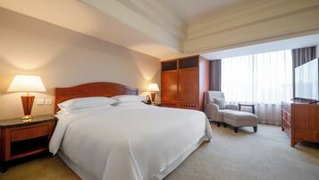 A hotel room with a large bed, two bedside tables with lamps, a wooden wardrobe, a seating area with a chair and a small table, and a television on a wooden cabinet.