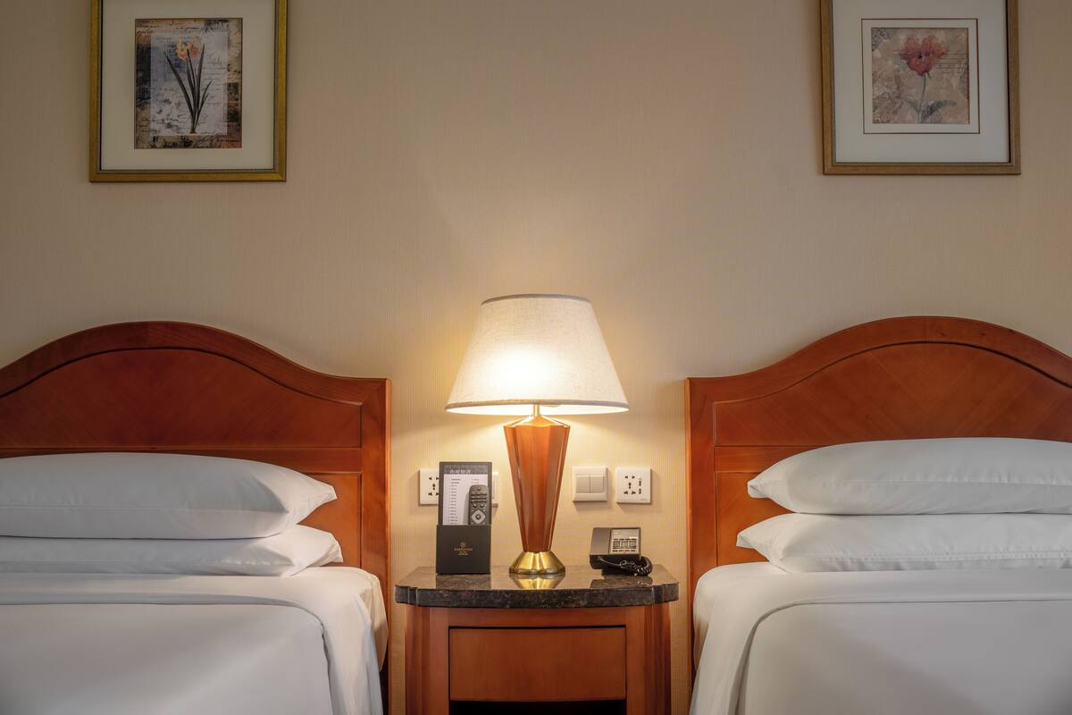 A hotel room with two beds, a nightstand with a lamp, a phone, and a remote control. There are framed pictures on the wall.