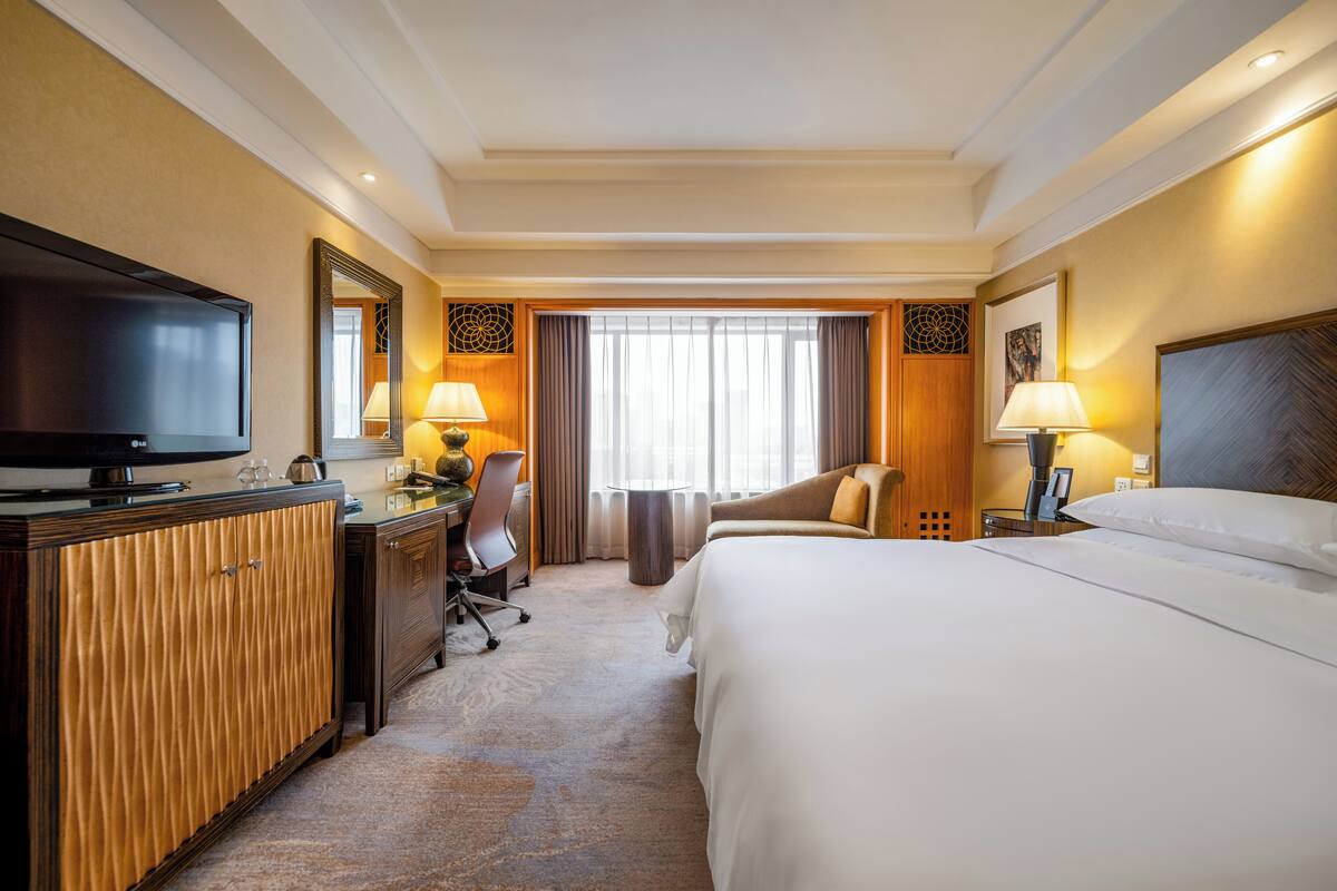A hotel room with a bed, a desk with a TV, a chair, a lamp, and a window with curtains.