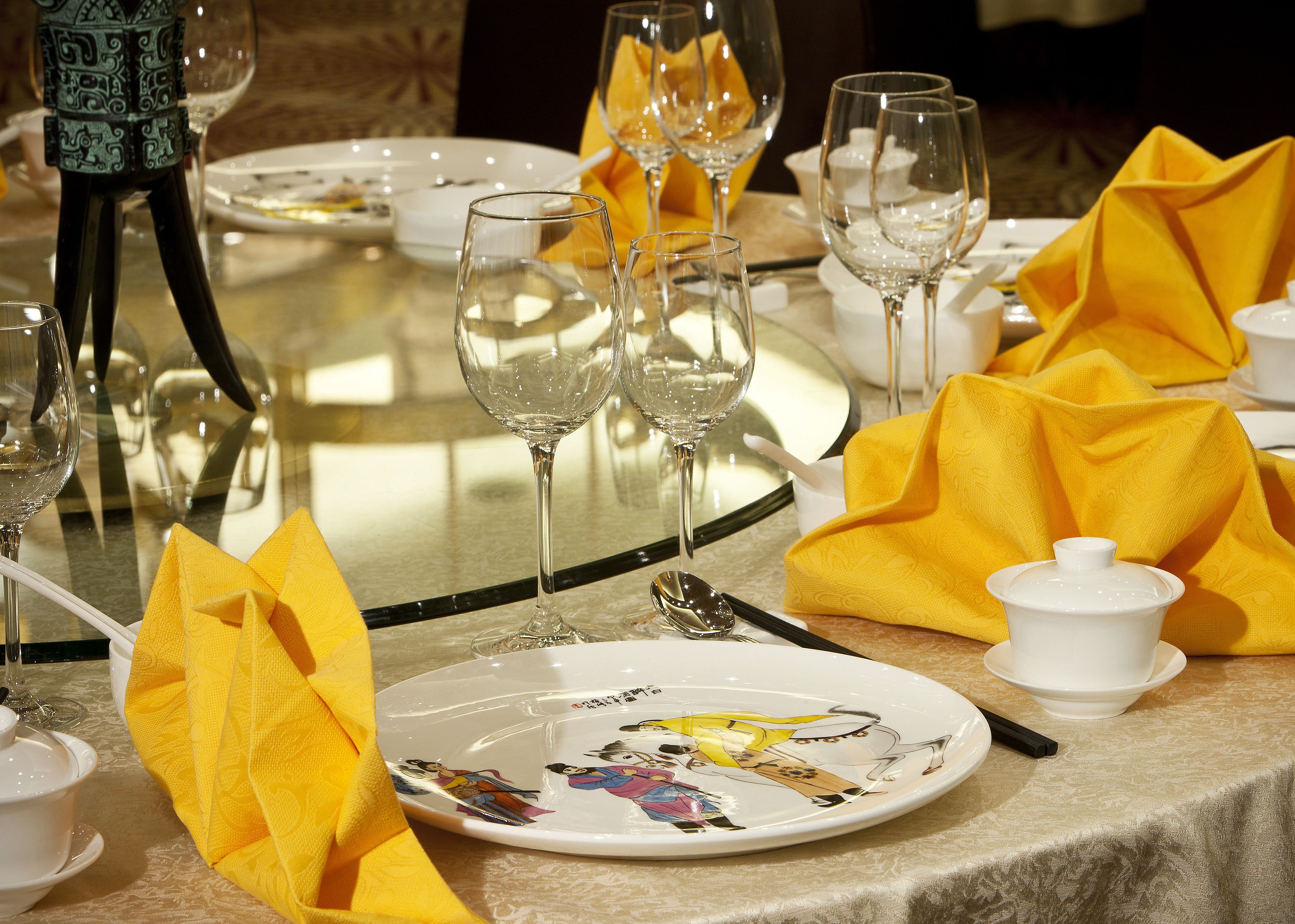 Una mesa lista para una comida, con varias copas de vino, un plato de diseño colorido y servilletas amarillas.