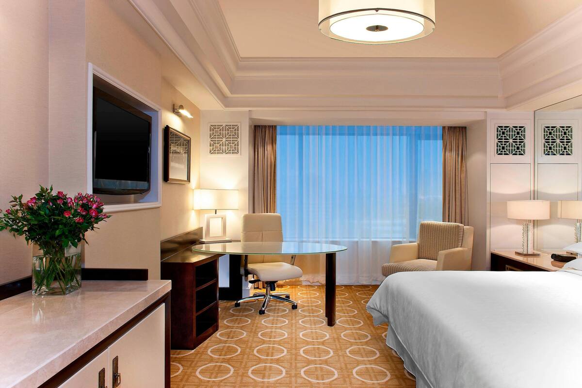 A hotel room with a bed, a desk with a chair, a TV, and a window with curtains.