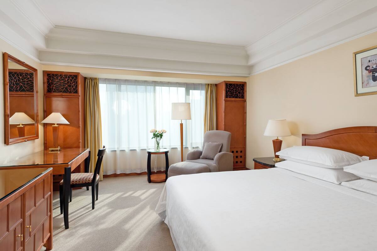 A hotel room with a bed, a desk, a chair, a small table with a vase, and a window with curtains.