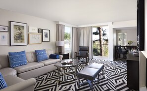 Un salon moderne avec un canapé, une table basse et un fauteuil. On peut voir des tableaux encadrés au mur, une fenêtre avec des rideaux et une vue sur les arbres à l’extérieur.