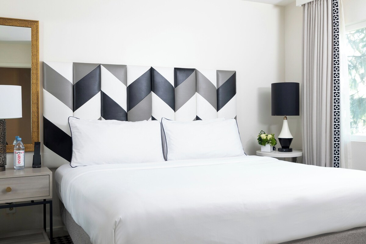 A hotel room with a bed, a nightstand, a lamp, a mirror, and a geometric wall art piece.