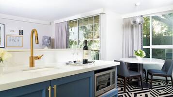 Une cuisine moderne avec un plan de travail blanc, un robinet doré et des armoires bleues. On y trouve un coin repas avec une table ronde et des chaises, ainsi qu’une grande fenêtre avec des rideaux.