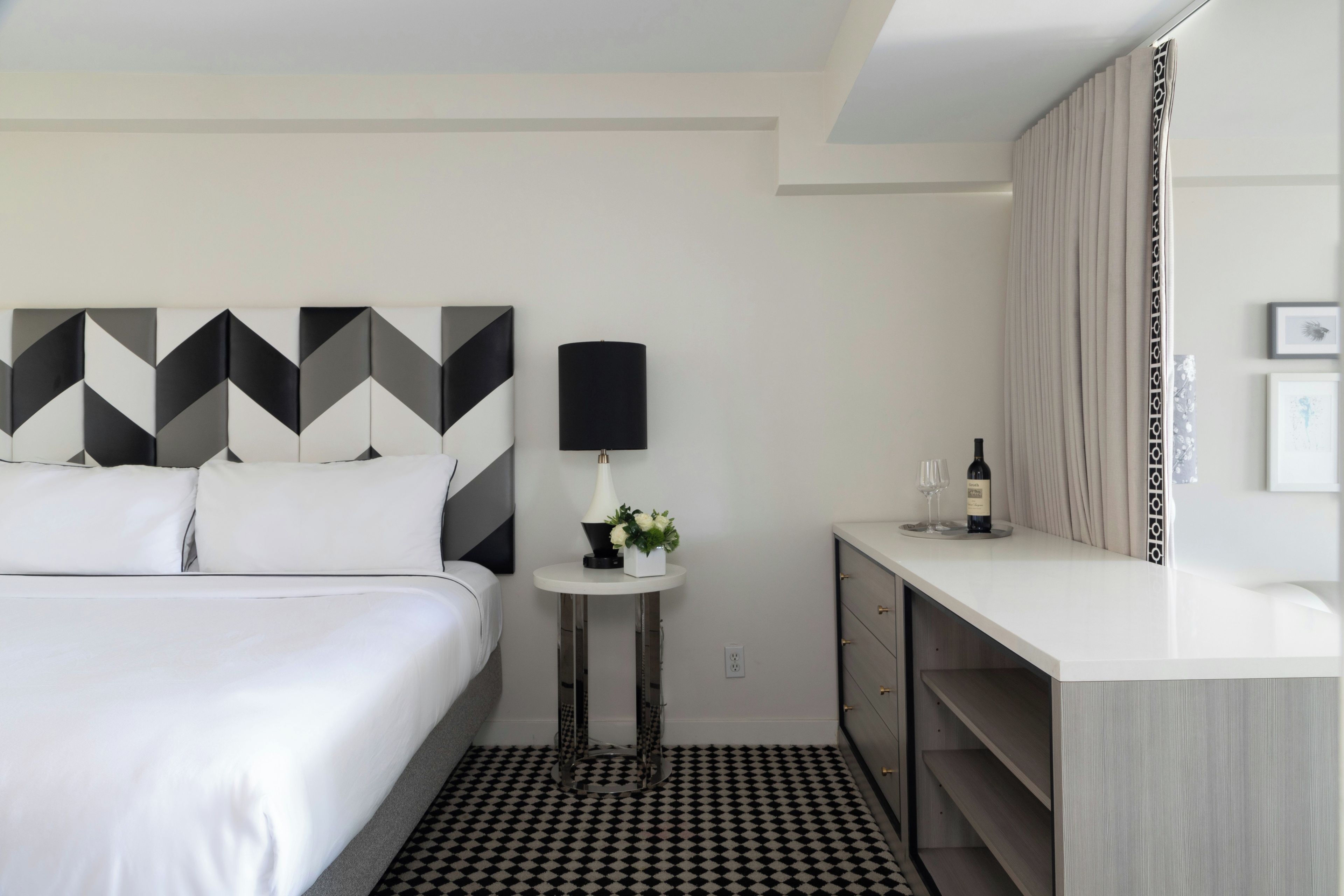 A hotel room with a bed, a small round table with a lamp, a wine bottle, and a mirror.