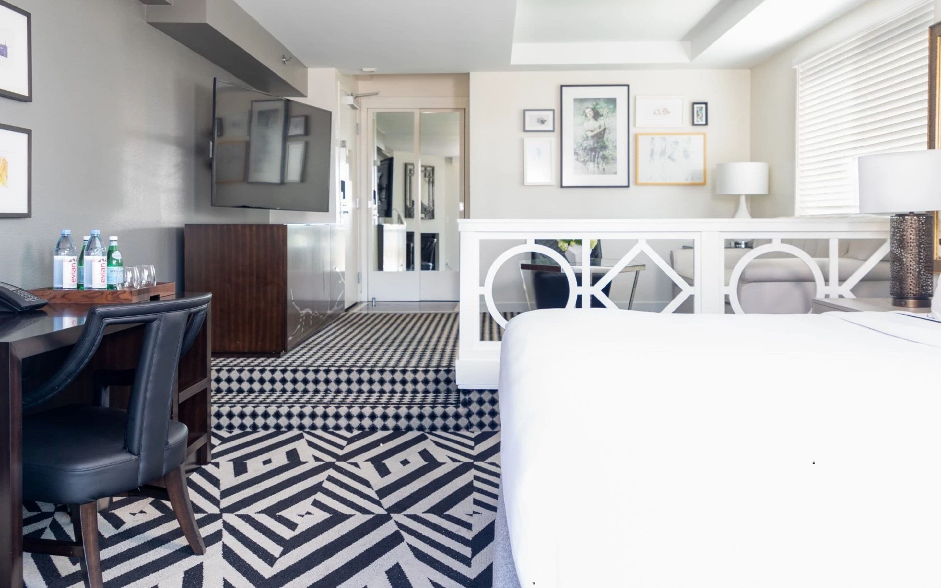 Una habitación de hotel moderna con suelo estampado en blanco y negro, un escritorio con silla, un televisor montado en la pared y una cama blanca.