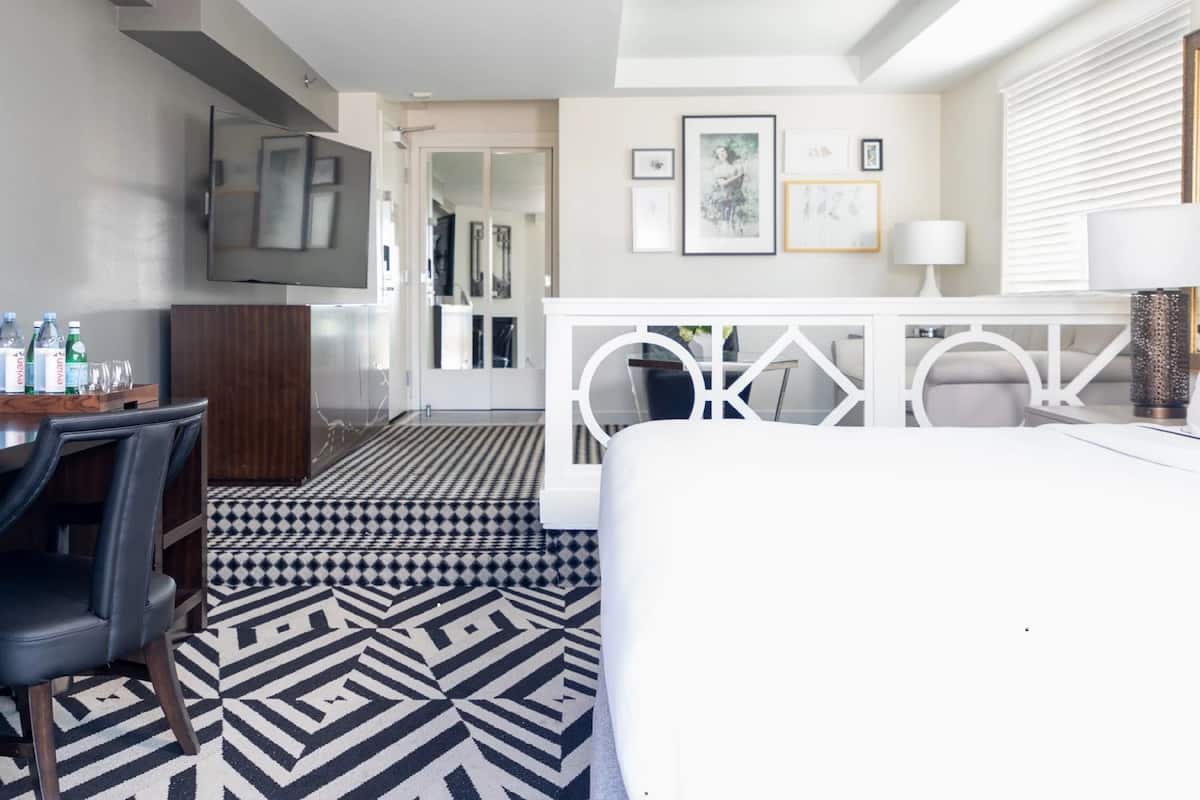 A modern hotel room with a black and white patterned floor, a desk with a chair, a television mounted on the wall, and a white bed.