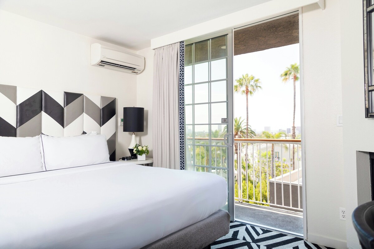 A hotel room with a bed, a nightstand, a lamp, a window with a view of palm trees, and a balcony.